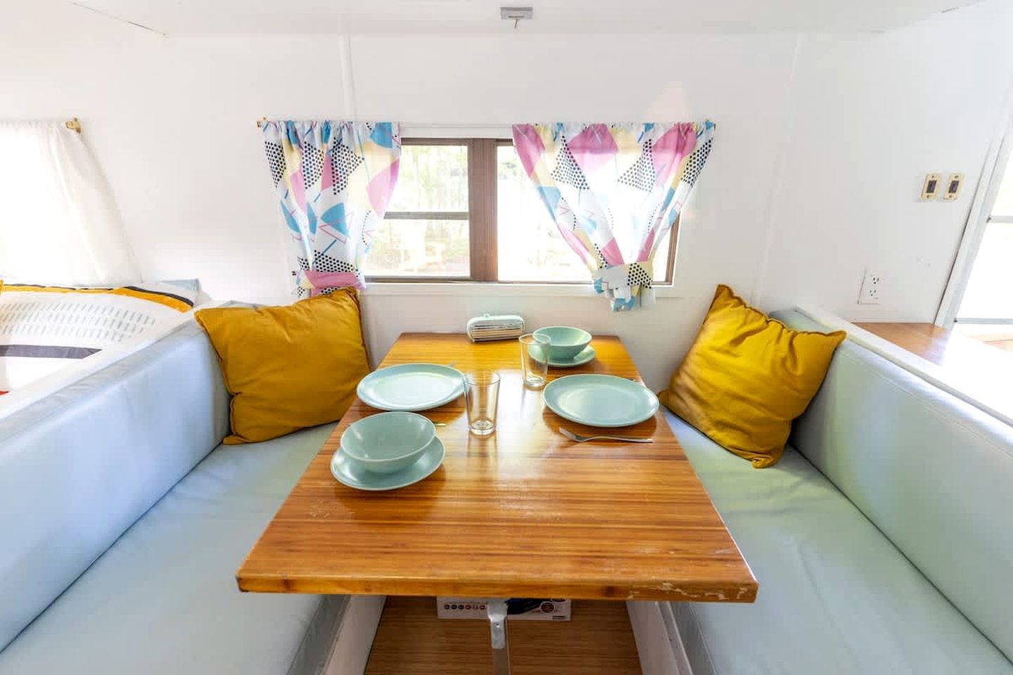 Un espace de vie compact comprenant une table en bois dressée pour les repas, deux coins salon avec des sièges confortables et une fenêtre agrémentée de rideaux à motifs.