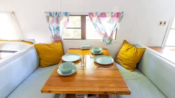 Un espace de vie compact comprenant une table en bois dressée pour les repas, deux coins salon avec des sièges confortables et une fenêtre agrémentée de rideaux à motifs.