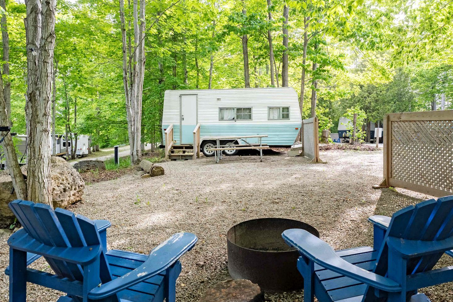 Un terrain de camping avec une caravane, deux chaises bleues et un foyer.