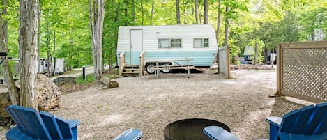 Un camping con una caravana, dos sillas azules y un hogar para la fogata.