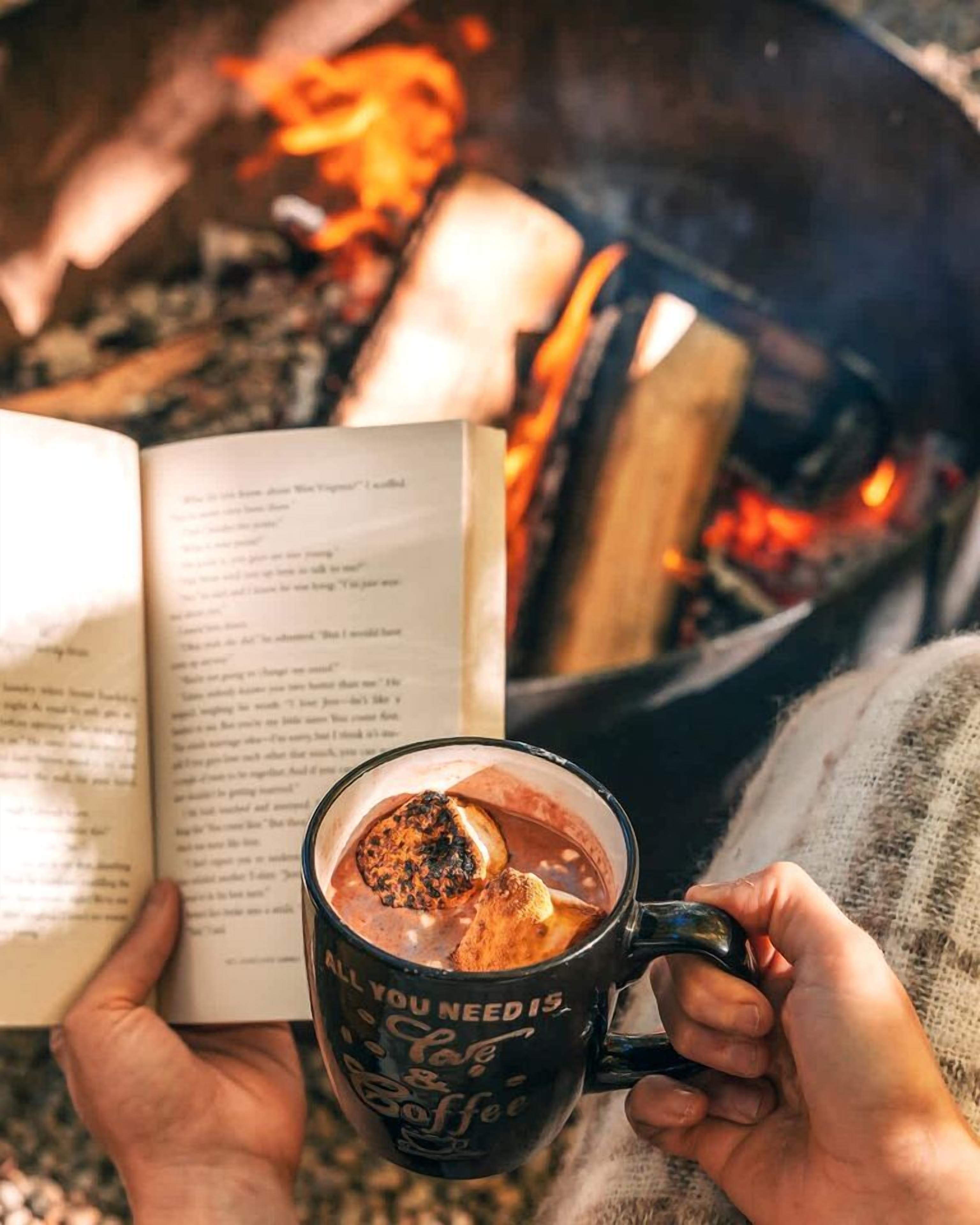 Une personne tenant un livre et une tasse contenant une boisson chaude, avec un feu en arrière-plan.