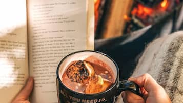 Une personne tenant un livre et une tasse contenant une boisson chaude, avec un feu en arrière-plan.