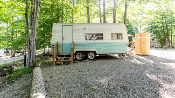 A vintage camper parked in a wooded area with a gravel ground, surrounded by trees.