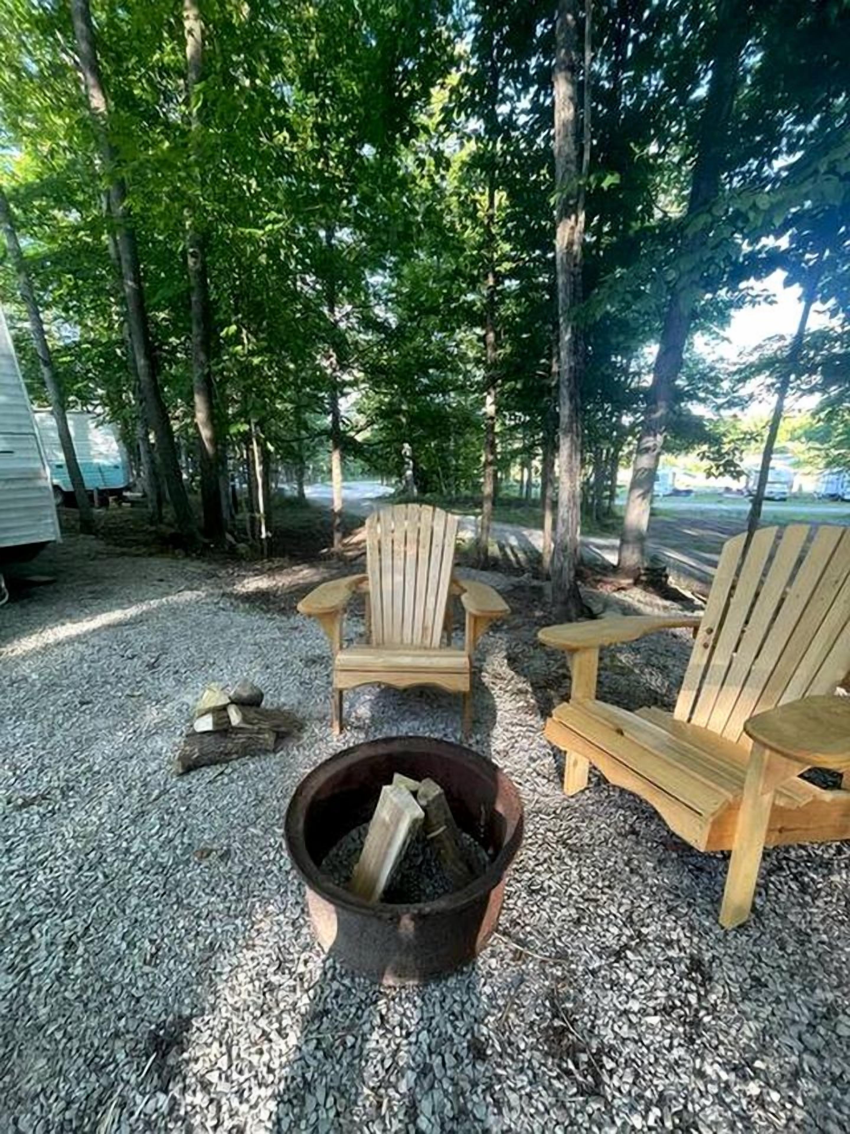 Un terrain de camping avec deux chaises Adirondack, un foyer et des arbres en arrière-plan.