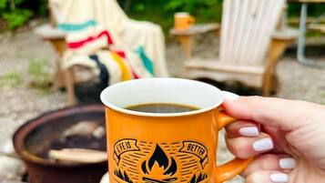 A person holding a mug with a campfire and chairs in a forest setting.
