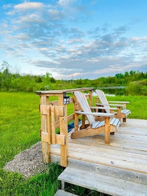 Eine Holzterrasse mit Adirondack-Stühlen und einem kleinen Tisch, mit Blick auf ein Grasfeld und ein Gewässer.
