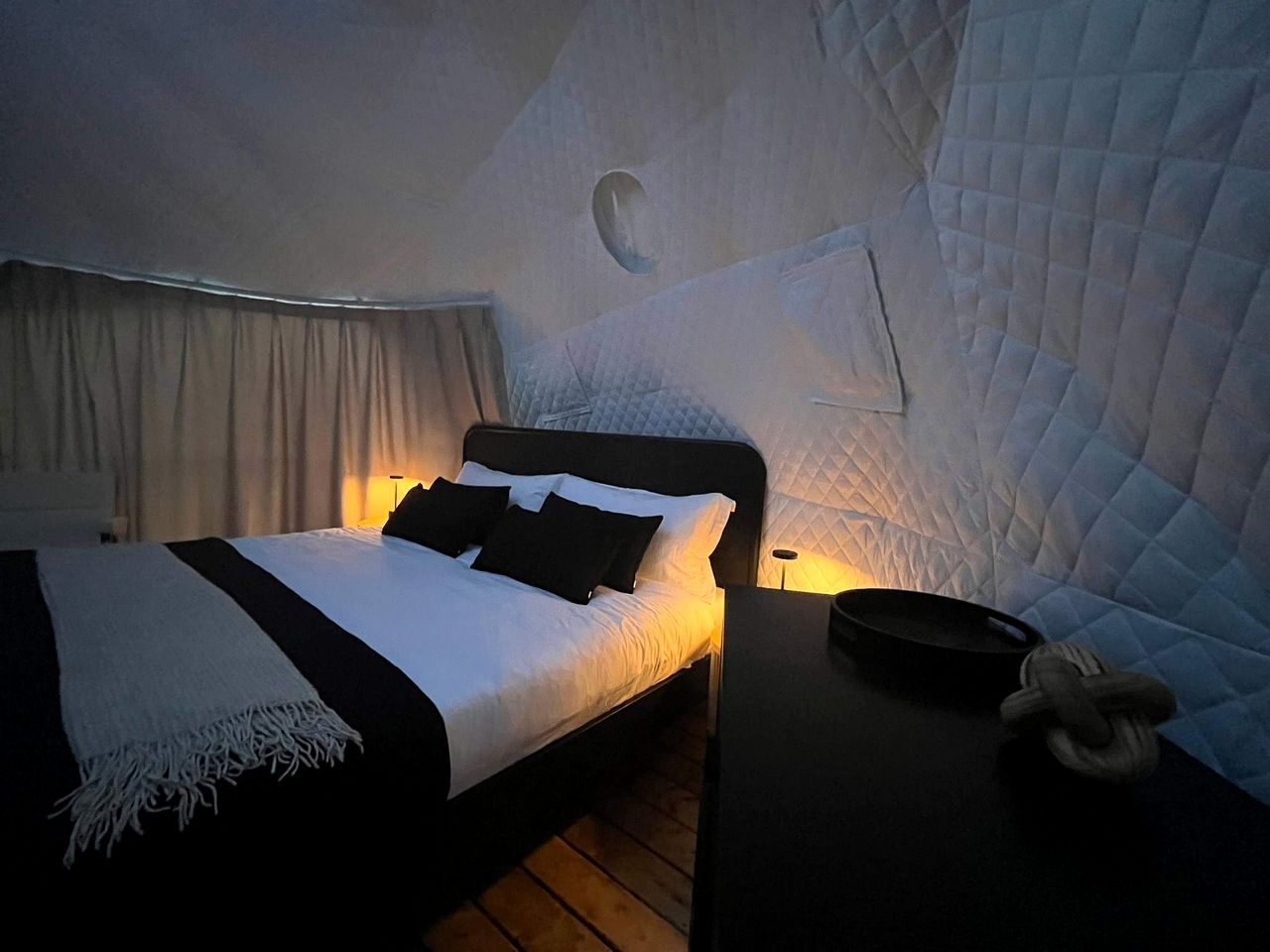 A bed with white bedding and black pillows, a bedside table with a lamp, and a quilted wall.