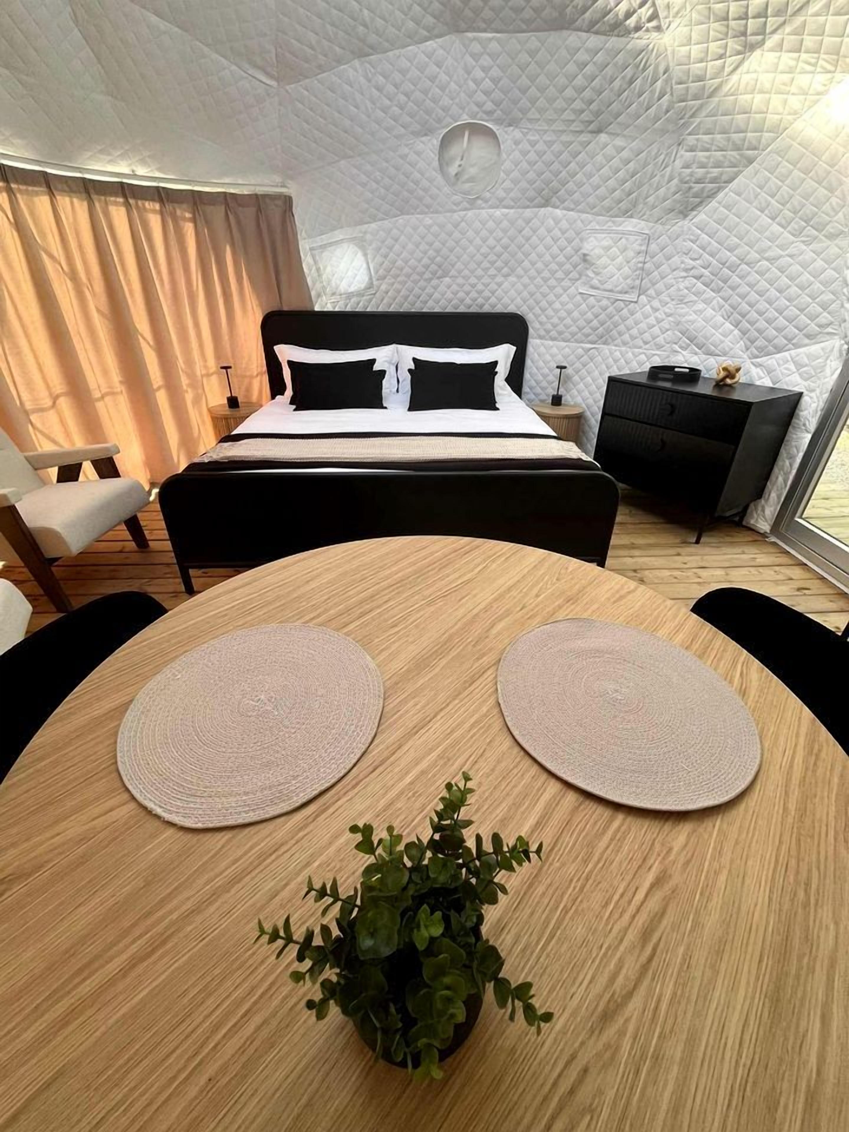 A bedroom with a bed, bedside tables, a small desk, a round wooden table with placemats, and a potted plant.