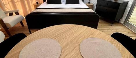 A bedroom with a bed, bedside tables, a small desk, a round wooden table with placemats, and a potted plant.