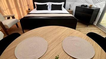 A bedroom with a bed, bedside tables, a small desk, a round wooden table with placemats, and a potted plant.