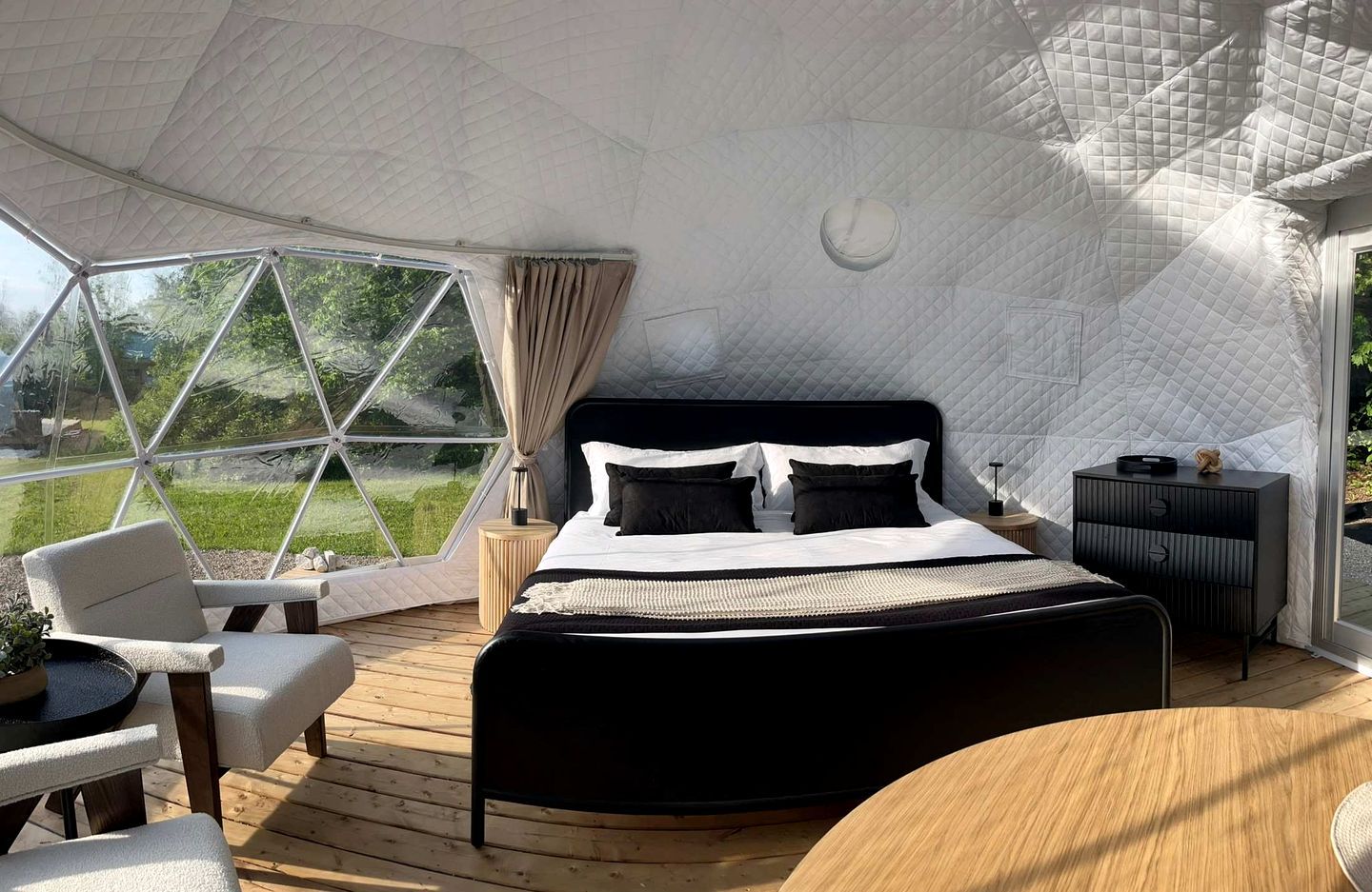 Una habitación amplia con una cama grande, suelo de madera, una mesa redonda y vistas a través de un ventanal.