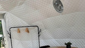 Una habitación con carpa, una mesa redonda de madera, sillas negras, un ventilador y un perchero metálico negro con ganchos.