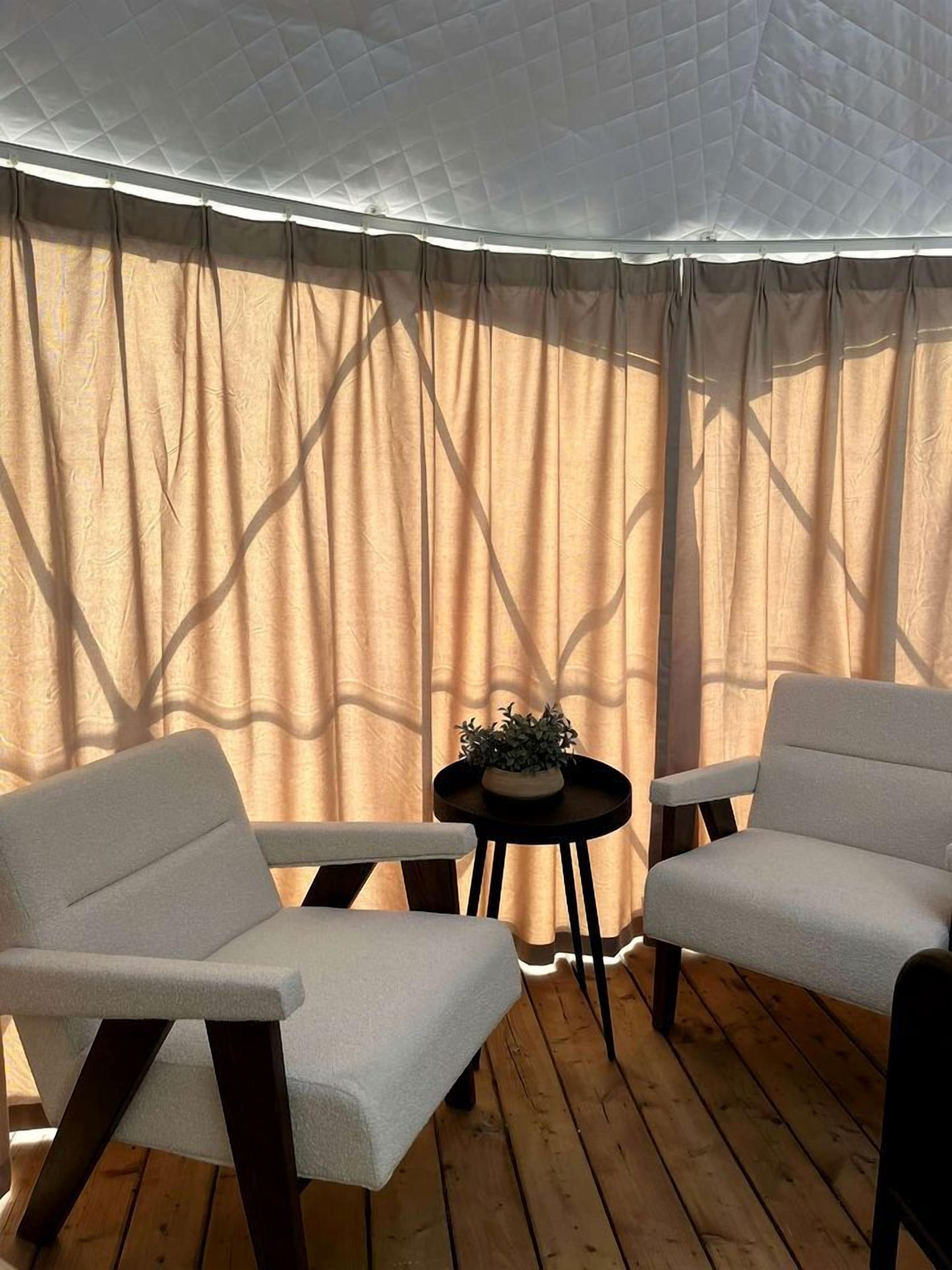 A room with a wooden floor, two white armchairs, a small round table with a plant, and sheer curtains.
