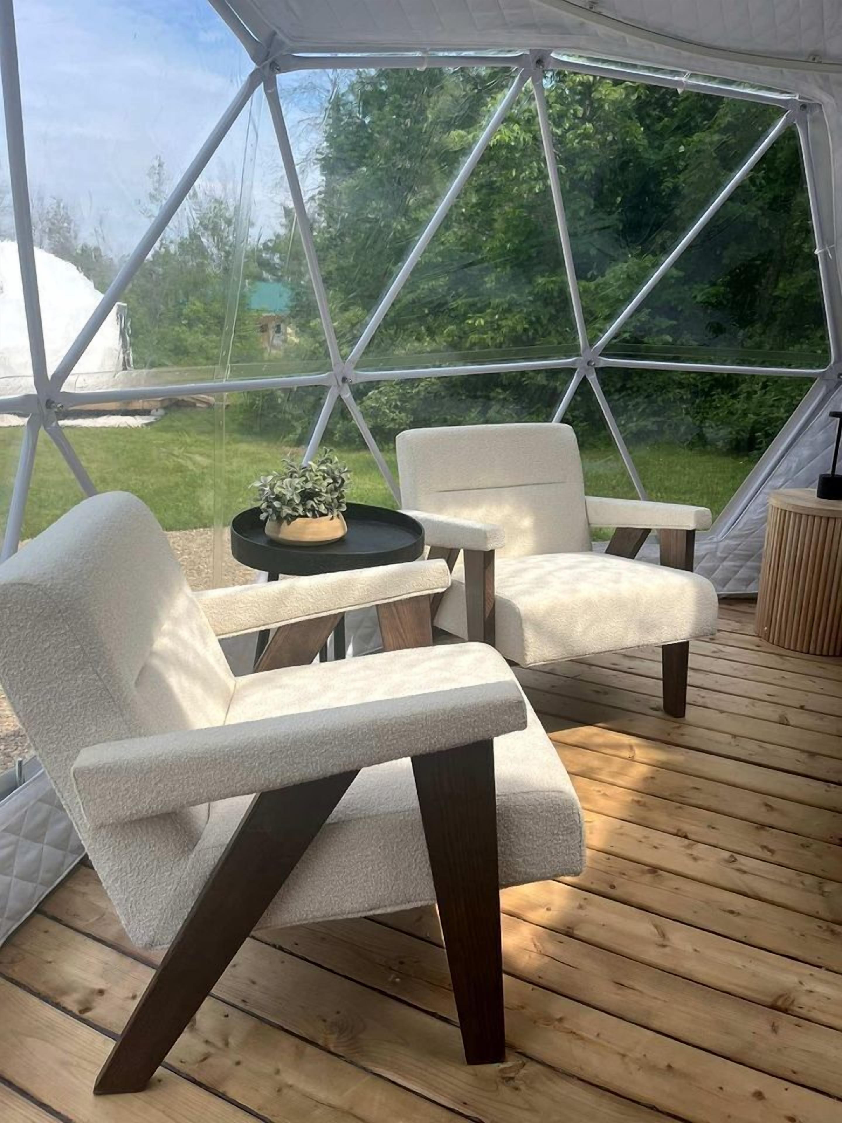 A tent with a wooden deck, two white cushioned chairs, a small table with a plant, and a view of greenery outside.