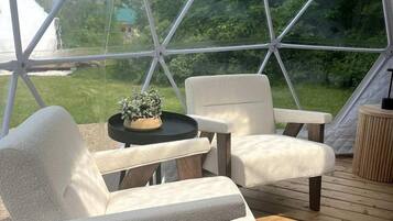 Ein Zelt mit einer Holzterrasse, zwei weißen gepolsterten Stühlen, einem kleinen Tisch mit einer Pflanze und Blick ins Grüne.