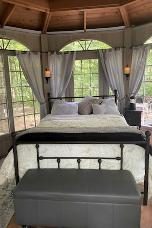 A bed with a headboard, a bedside table with a lamp, and curtains on large windows.