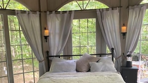 A bed with a headboard, a bedside table with a lamp, and curtains on large windows.