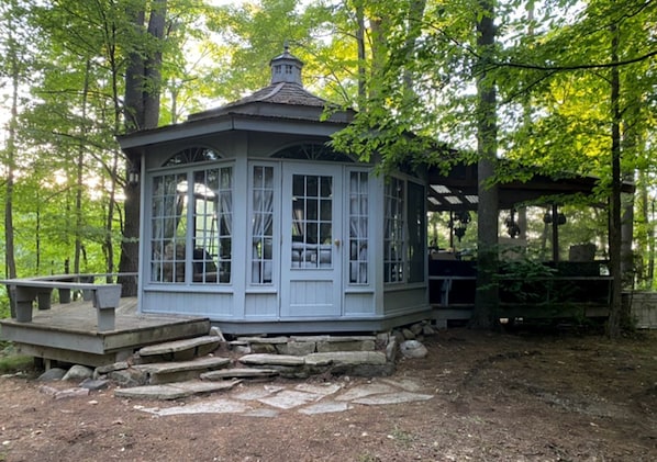 A gazebo with a stone foundation, large windows, and a roof with a chimney.