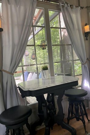 A room with a table and two stools, a window with curtains, and a hanging light fixture.