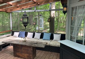 A covered outdoor seating area with a wooden table, cushioned benches, and hanging lanterns.