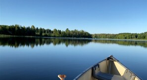 Un lago tranquilo con un bote en primer plano y una orilla boscosa al fondo.