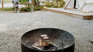 Ein runder Grill für den Außenbereich mit einem Grillsteak, umgeben von Kies, mit einem Zelt und einer Person im Hintergrund.