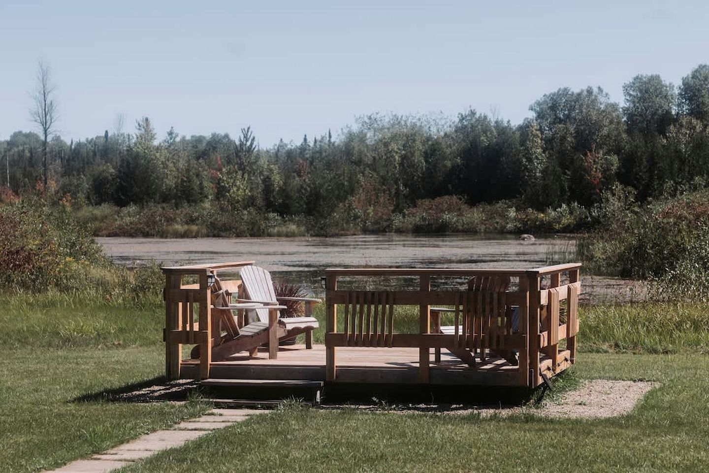 A wooden deck with chairs and a table, surrounded by a grassy area and a forested backdrop.