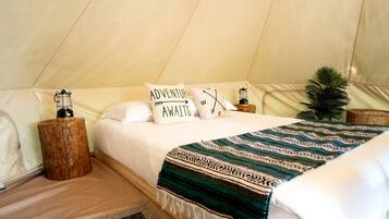 A tented accommodation room with a bed, pillows, a blanket, a lantern, and a small plant.