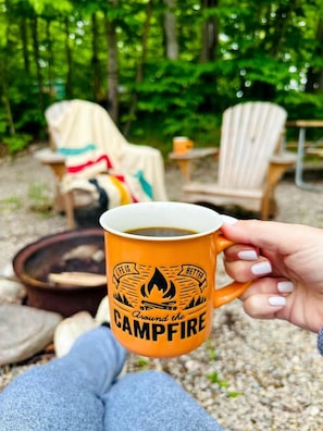 A person holding a mug with a campfire and chairs in a forest.