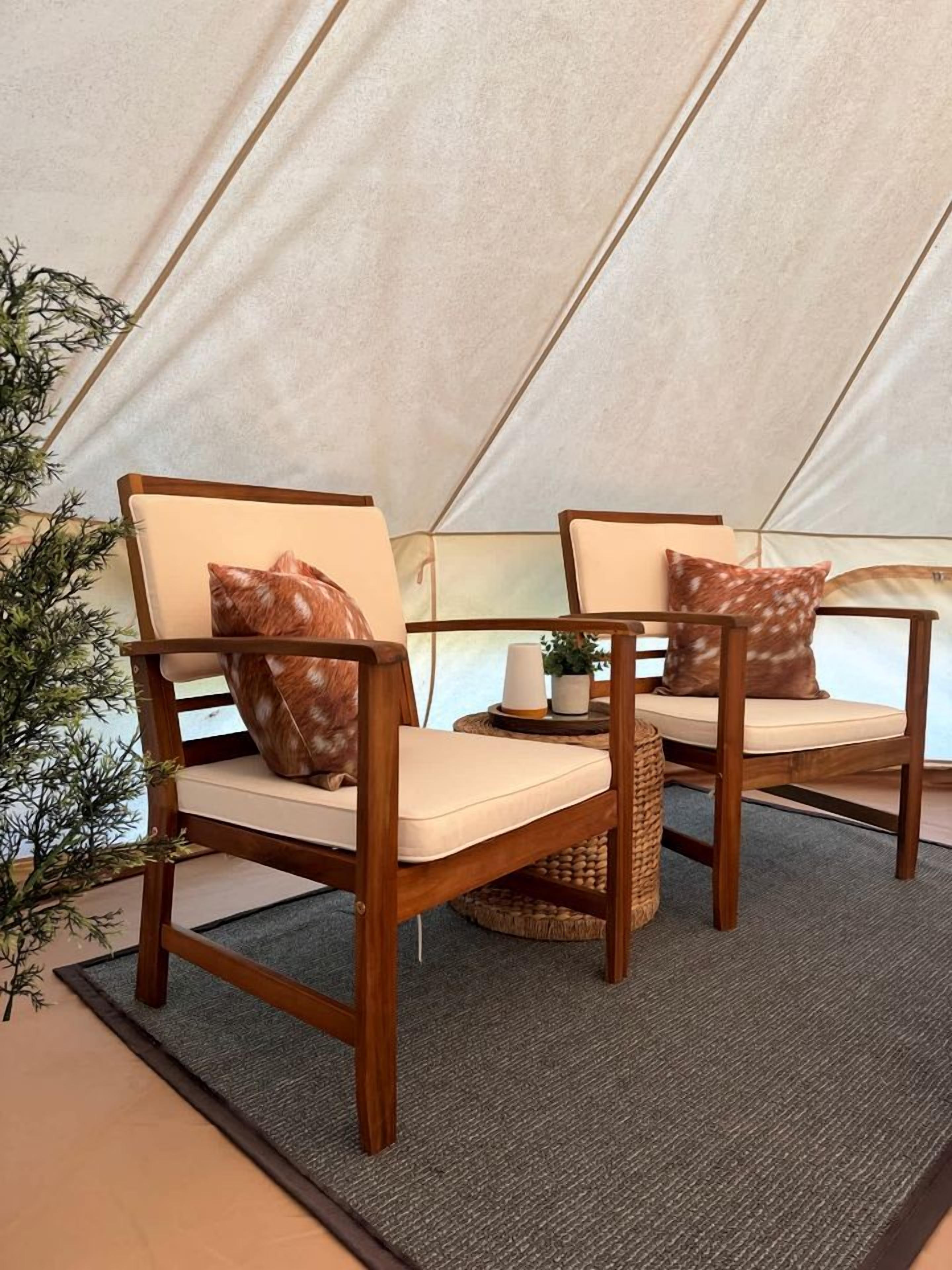 Une tente avec deux chaises en bois, chacune agrémentée d’un coussin et d’un oreiller à motifs, ainsi qu’une petite table sur laquelle sont posés une bougie et une plante.