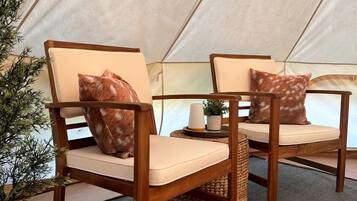 A tent with two wooden chairs, each with a cushion and a patterned pillow, and a small table with a candle and a plant.