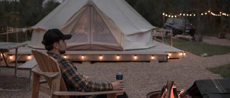 A person seated at a table with a campfire, a tent, and string lights in the background.