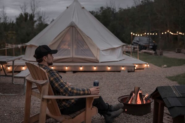 Une personne assise à une table, avec un feu de camp, une tente et des guirlandes lumineuses en arrière-plan.