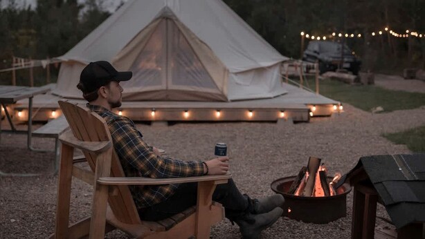 Eine Person sitzt an einem Tisch mit Lagerfeuer, Zelt und Lichterkette im Hintergrund.