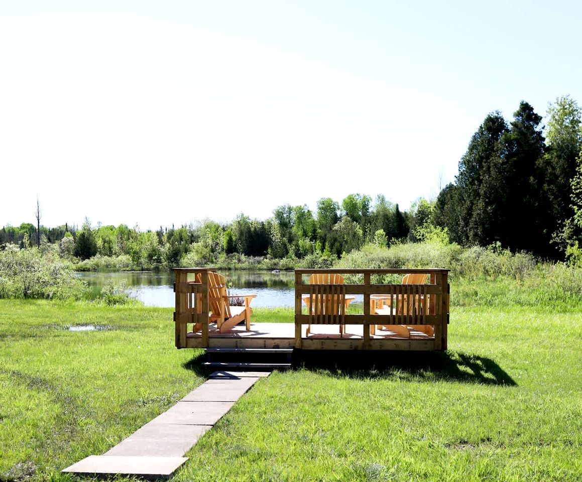 Una terraza de madera con sillas y una mesa, un área con césped y una masa de agua al fondo.