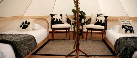 A cozy tented room with two beds, each with a black and white bear-themed pillow, a wooden chair, and a small table with a plant.
