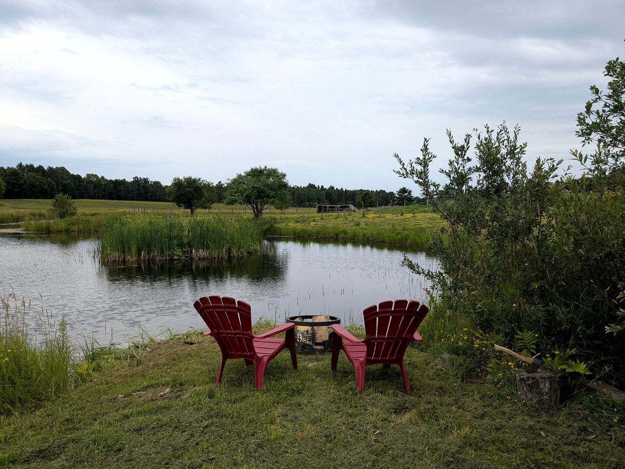 Twee Adirondack-stoelen gericht op een vijver met een vuurkuil in het midden.