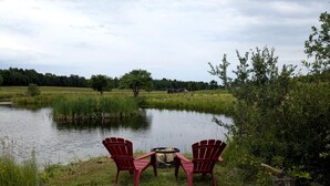 Two Adirondack chairs facing a pond with a fire pit in the center.