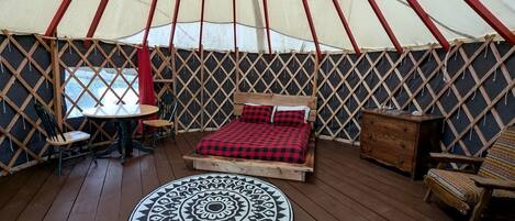 Ein Jurteninneres mit einem Bett, einem runden Teppich, einem Tisch, Stühlen und einer Holzkommode.