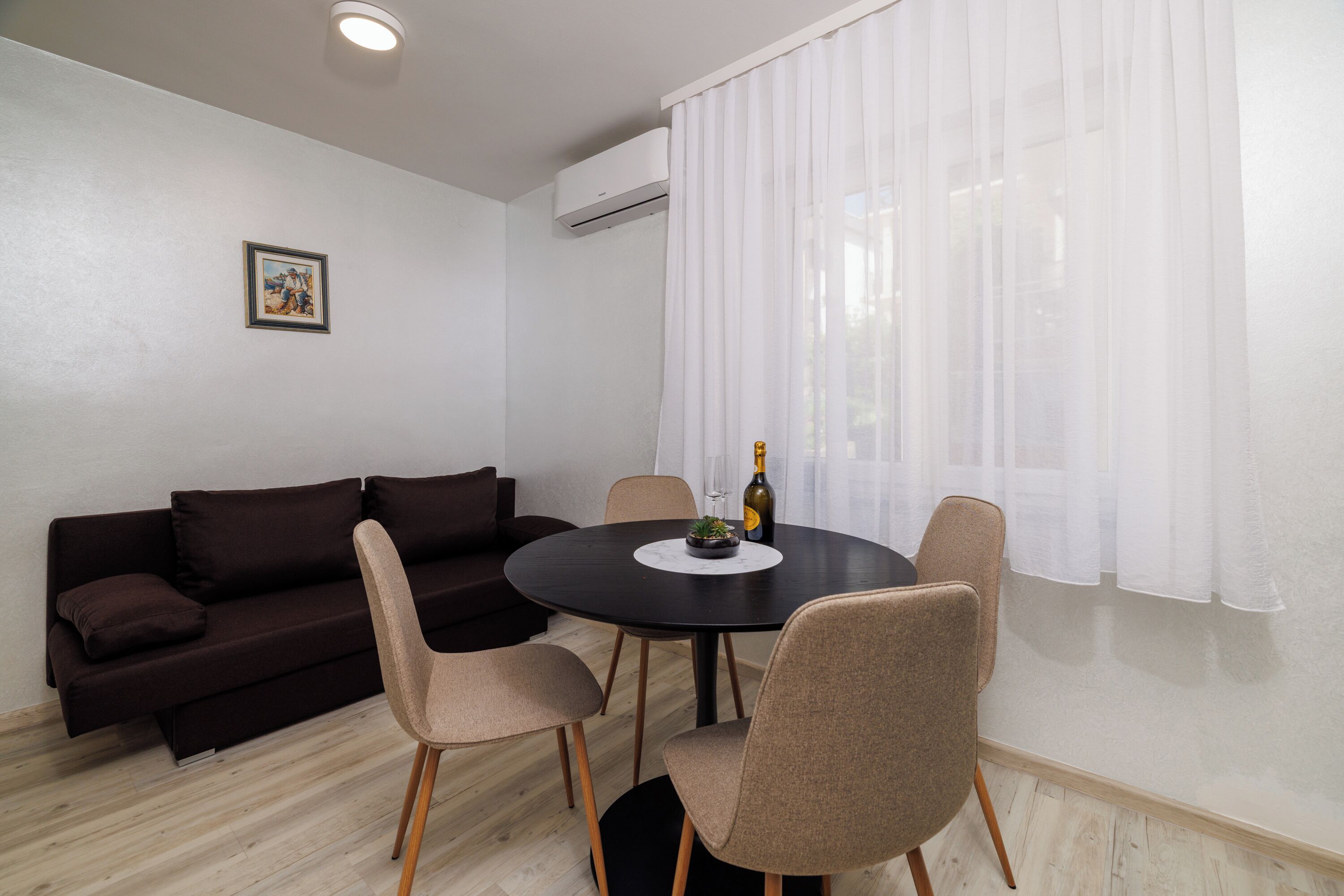 A room with a dark brown sofa, a round table with chairs, a bottle of champagne, and a picture on the wall.