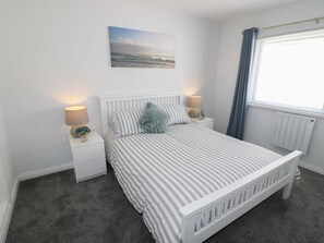 A bedroom with a bed, two bedside tables, a lamp, a radiator, and a window with curtains. - The Cartwheel, Amroth (Narberth)