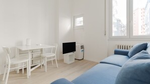 A room with a white desk, two chairs, a TV, a white air conditioner, a blue sofa, and a window with a view of buildings. - GuestHost - Comfortable 30m2 studio flat, perfect for 2 people, located on the mezzanine floor of a building (steps to access).The accommodation is located in the Calvairate district, a residential area characterized by the green heart of Piazza Insubria (Milan)