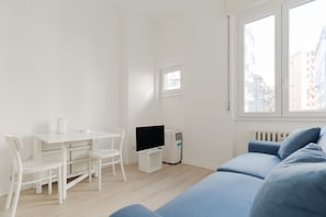 Quarto com escrivaninha branca, duas cadeiras, televisão, ar condicionado branco, sofá azul e janela com vista para edifícios.