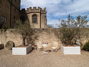 A courtyard with a stone building featuring a large arched window and a castle-like tower. There are two white planters with trees and a small table with chairs. - Snape Castle Under Croft and Gardens, Bedale (Bedale)