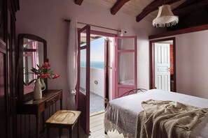 Ein Schlafzimmer mit Bett, Sessel, Schminktisch und Spiegel, mit Blick auf das Meer durch eine offene Tür.