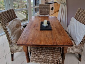 Una mesa de madera para comer con sillas tejidas, una vela y vista a un jardín a través de un ventanal.