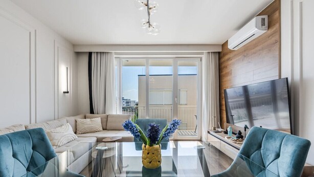 Un salon moderne avec un canapé, une table basse en verre, deux fauteuils bleus, un téléviseur et un balcon avec vue.