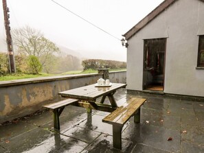 A wet outdoor patio with a picnic table and benches, a white building with a door, and a view of trees and hills in the background. - Penlon, Capel Bangor (Aberystwyth)