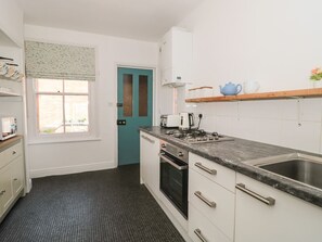 A kitchen with a window, a teal door, a stove, a sink, and a wooden shelf with a teapot. - Bluebell Apartment, Swanage (Swanage)