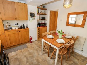 A kitchen with wooden cabinets, a dining table set for four, and a small side table with a vase of flowers. - Serenity Cottage, Oxenhope (Keighley)